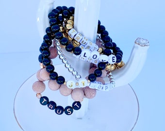 Può includere: Un set di cinque braccialetti con perline di diversi colori e ciondoli con lettere. I braccialetti sono esposti su un supporto a cactus in ceramica bianca. I ciondoli compongono le parole "TRUST", "LOVE", "HOPE", "JOY" e "JUST".