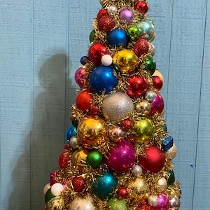 以下が含まれることがあります： 金色のティンセルで作られた円錐形の卓上クリスマスツリーの装飾品。赤、青、緑、ピンク、銀、金の様々な色合いの、光沢のあるキラキラした球形のオーナメントが密集して飾られています。