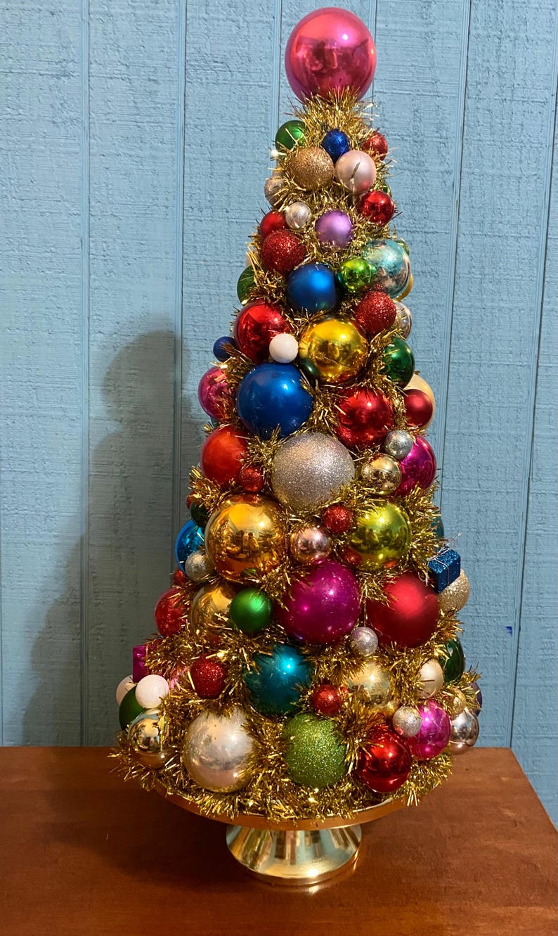 以下が含まれることがあります： 金色のティンセルで作られた円錐形の卓上クリスマスツリーの装飾品。赤、青、緑、ピンク、銀、金の様々な色合いの、光沢のあるキラキラした球形のオーナメントが密集して飾られています。