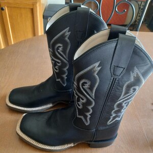 May include: A pair of black cowboy boots with white decorative stitching. The boots have a square toe and a pull-on loop at the top. The stitching forms a decorative pattern on the boot shaft.