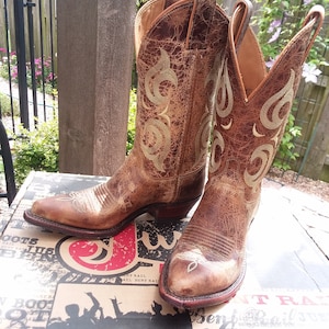 May include: A pair of brown leather cowboy boots with intricate gold embroidery. The boots are sitting on a box with the Justin Boots logo.