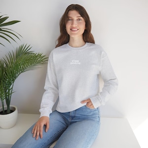 Könnte beinhalten: Ein hellgraues Sweatshirt mit dem Text "sleep pending" in Weiß. Das langärmlige Rundhals-Shirt ist aus einem weichen Material gefertigt. Das Model trägt eine blaue Jeans.
