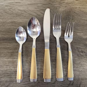 May include: A set of five pieces of silverware, including a spoon, a tablespoon, a knife, and two forks. The handles are gold-colored with a silver band at the base. The utensils are arranged on a wooden surface.