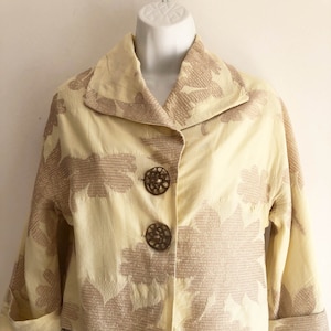 Può includere: Una giacca gialla corta con un motivo floreale marrone e tre bottoni decorativi. La giacca ha un colletto e maniche lunghe.