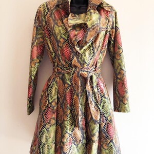 Könnte beinhalten: Ein langärmliger Trenchcoat mit Schlangenmuster in Grün-, Rot- und Brauntönen. Der Mantel hat einen Gürtel in der Taille, einen gerüschten Kragen und einen Knopfverschluss. Der Mantel wird auf einer schwarzen Schaufensterpuppe präsentiert.