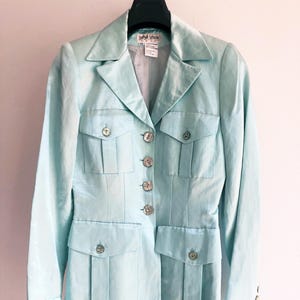 Puede incluir: Una blazer azul claro con cuatro bolsillos delanteros y botones plateados. La blazer tiene un cuello clásico y mangas largas. La etiqueta dice "Sophie Sitron". La chaqueta está colgada en una percha negra.