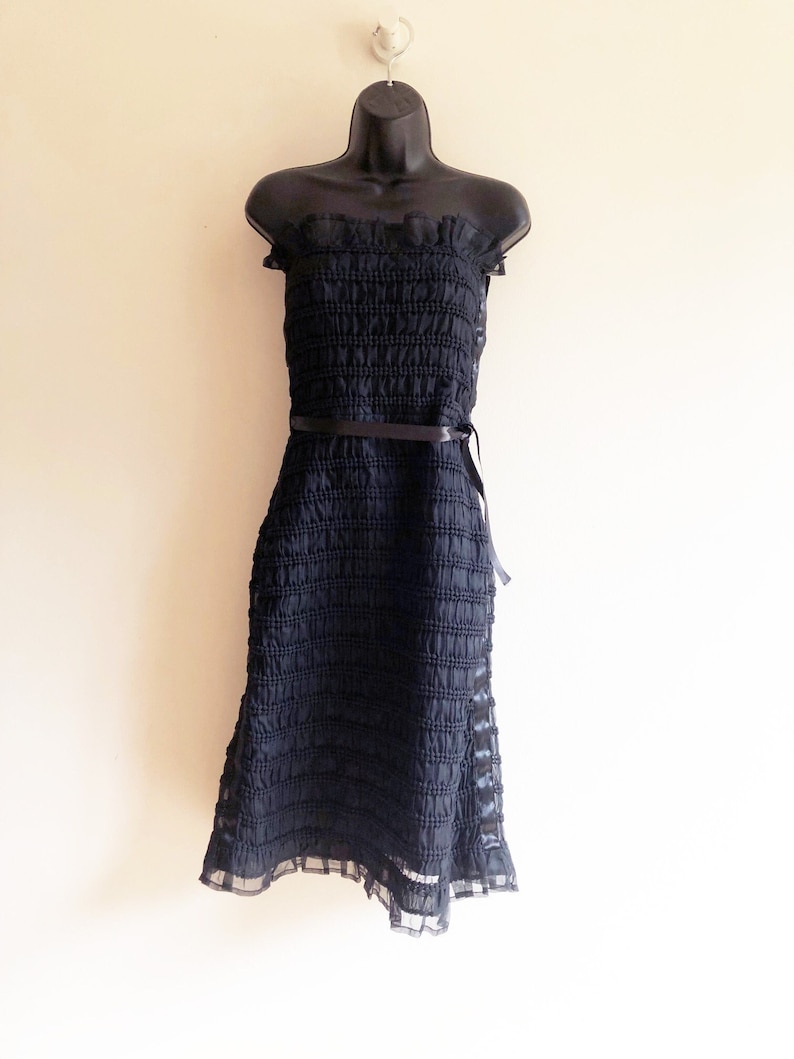 May include: A black strapless dress with a ruffled neckline and a black ribbon belt. The dress is made of a textured fabric and has a flowing skirt.