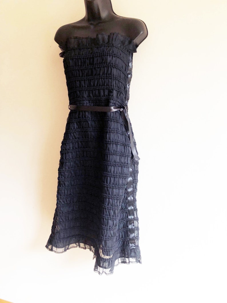 May include: A black strapless dress with a ruffled neckline and a black ribbon belt. The dress is made of a textured fabric and has a flowing skirt.