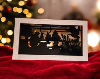 May include: A framed silhouette artwork showing a hunter and dog in a forest setting. The black cut-out is set against a warm, glowing backdrop, within a light-coloured wooden frame. The piece is displayed on a red velvet surface.