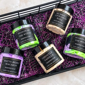May include: Five clear plastic jars with black lids containing whipped soap and body butter. Labels read: Batty Berry Macaron, Season of the Witch, Mummy's Cinnamon Roll, and Ghostly Vanilla, all by Sassy Girl Beautique. The jars feature colorful creams.