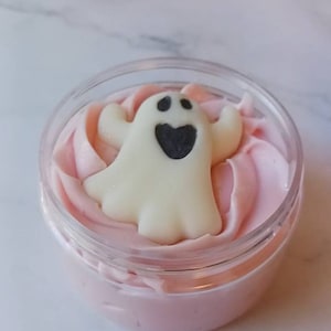 May include: A clear plastic jar filled with light pink whipped cream or body butter. Resting on top of the pink cream is a small, off-white ghost-shaped decoration with two black eyes and a black heart-shaped mouth, centered in the container.