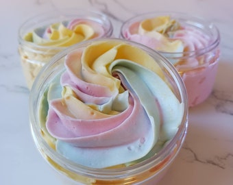 May include: Three clear plastic jars filled with colorful, swirled whipped cream. The colors include pastel yellow, pink, blue, and peach. The jars are arranged on a white marble surface.