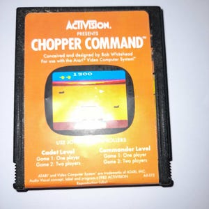 May include: A black Atari video game cartridge for Chopper Command by Activision. The orange label features the game title, a screenshot of the gameplay showing a score of 1300, and text detailing game levels for one or two players.