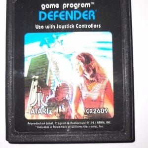 Puede incluir: Un cartucho de videojuego Atari negro y rectangular para el juego Defender. La etiqueta presenta el texto game program DEFENDER, Use with Joystick Controllers, el logotipo de Atari y CX2609. La ilustración muestra una persona y una ciudad.