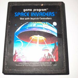 Peut inclure: Une cartouche de jeu vidéo Atari 2600 rectangulaire noire pour Space Invaders. L'étiquette présente une illustration bleue et blanche d'une soucoupe volante au-dessus d'un paysage rouge, avec le texte game program, SPACE INVADERS et CX2632.
