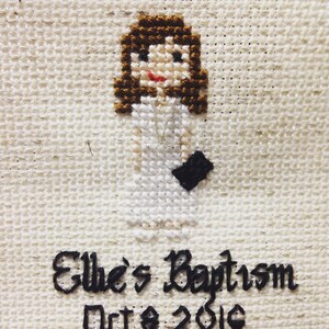 May include: Cross-stitch embroidery of a girl in a white dress with brown hair. The text below reads "Elle's Baptism Oct. 2016".