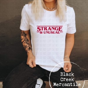 May include: White t-shirt with a red graphic that says "STRANGE -& UNUSUAL-." The graphic is in a retro font style. The t-shirt is worn by a person with a tattoo on their arm. The text "Black Creek Mercantile" is visible in the bottom right corner of the image.