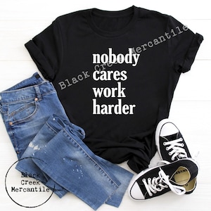 May include: Black t-shirt with white text that reads "nobody cares work harder". The shirt is displayed on a white surface with blue jeans and black sneakers.
