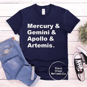 May include: Navy blue t-shirt with white text: Mercury & Gemini & Apollo & Artemis. The shirt is paired with blue jeans and black and white sneakers. The Black Creek Mercantile logo is visible.