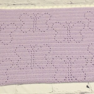 May include: A handmade, lavender crochet blanket with a white scalloped border. The blanket features a repeating pattern of openwork hearts. This soft, cozy blanket is perfect for a baby or child.