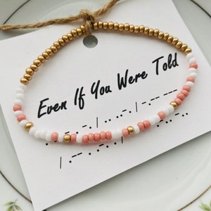 May include: A beaded bracelet with white and pink beads and gold accents. The bracelet has the text "Even If You Were Told" and braille on a white card.