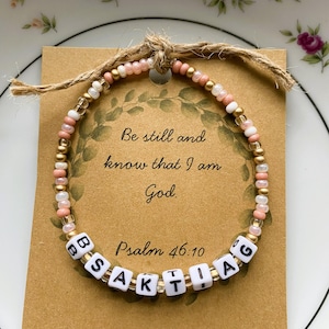 May include: A beaded bracelet with a message card. The bracelet features gold, white, and pink beads. The card reads "Be still and know that I am God. Psalm 46:10". The bracelet is tied with twine.