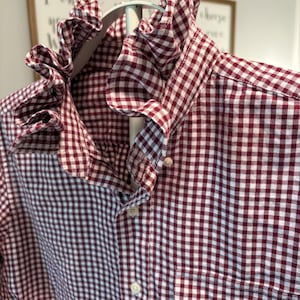 May include: A burgundy and white gingham patterned shirt with a ruffled collar and button-down front. The shirt is hanging on a white hanger, showcasing the classic check design. The fabric appears to be a lightweight cotton or similar material.