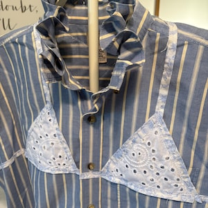 May include: A blue and white striped button-down shirt featuring a ruffled collar and two white eyelet lace triangle patches stitched onto the front. The shirt has a front button placket with brown buttons and a visible interior garment tag.