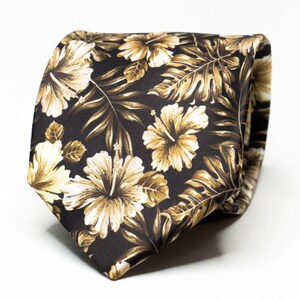 Könnte beinhalten: Eine braune Seidenkrawatte mit einem floralen Muster aus weißen und goldenen Hibiskusblüten und Blättern.