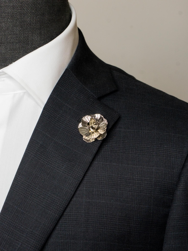 May include: A silver flower lapel pin is attached to a dark grey plaid suit jacket. The pin features a detailed floral design, adding a touch of elegance. The suit is paired with a white dress shirt and a crisp white collar.