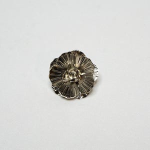 May include: A silver-coloured, flower-shaped brooch. The brooch features multiple layers of petals radiating from a central point. The petals have a textured, ridged surface, giving the brooch a detailed appearance. The brooch is set against a white background.