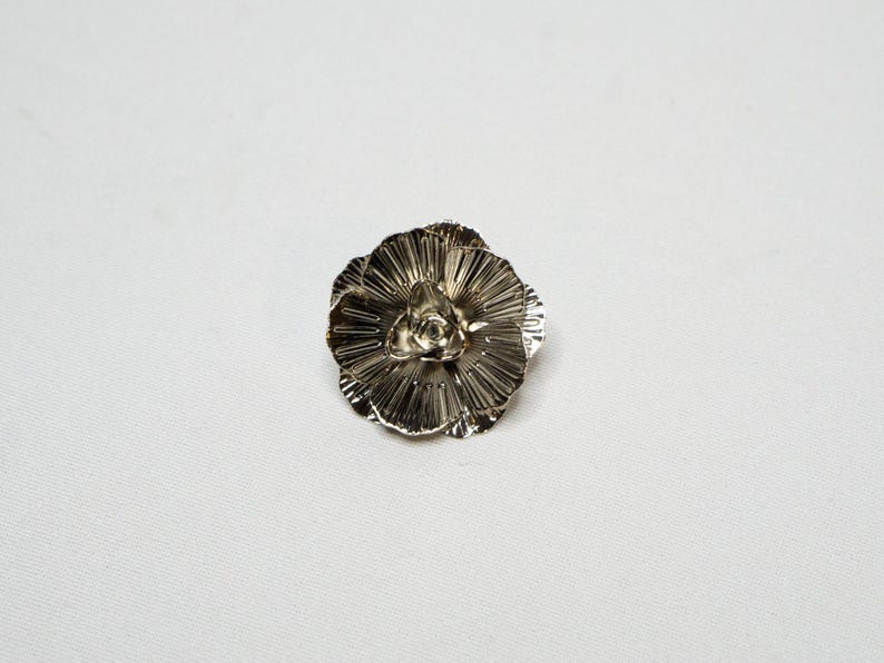 May include: A silver-coloured, flower-shaped brooch. The brooch features multiple layers of petals radiating from a central point. The petals have a textured, ridged surface, giving the brooch a detailed appearance. The brooch is set against a white background.