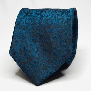 Pode incluir: Uma gravata azul-petróleo com um padrão floral escuro. A gravata é feita de um tecido texturizado e está atada, mostrando sua ponta. Um acessório clássico para trajes formais ou de negócios.