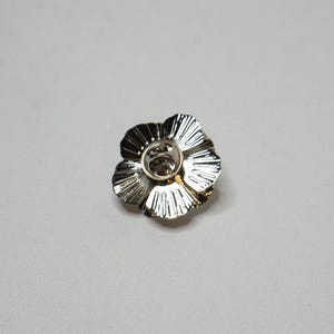 May include: A silver flower-shaped brooch with a textured finish. The brooch features a central pin and clasp. The petals have a ridged design, adding detail. This decorative brooch is suitable for clothing or accessories.
