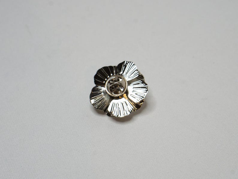 May include: A silver flower-shaped brooch with a textured finish. The brooch features a central pin and clasp. The petals have a ridged design, adding detail. This decorative brooch is suitable for clothing or accessories.