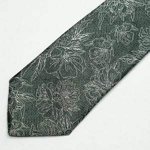 May include: A dark green necktie with a floral pattern. The tie features a repeating design of white flowers and leaves. The fabric has a textured appearance, and the tie is angled across a white surface.
