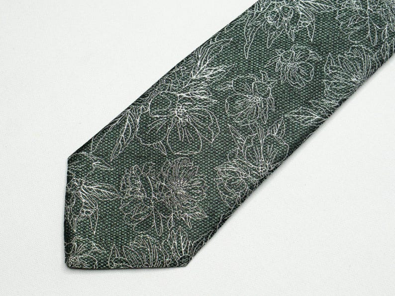 May include: A dark green necktie with a floral pattern. The tie features a repeating design of white flowers and leaves. The fabric has a textured appearance, and the tie is angled across a white surface.