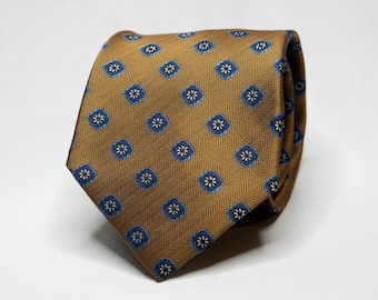May include: A folded tie with a repeating pattern of blue and white floral designs set in blue squares on a gold background. The tie has a herringbone weave pattern.