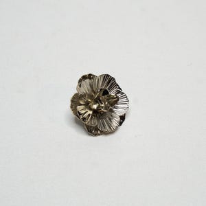 May include: A silver-coloured metal flower brooch. The brooch features multiple layers of petals with a textured, ridged design. The flower is centered on a white background, highlighting its intricate details.