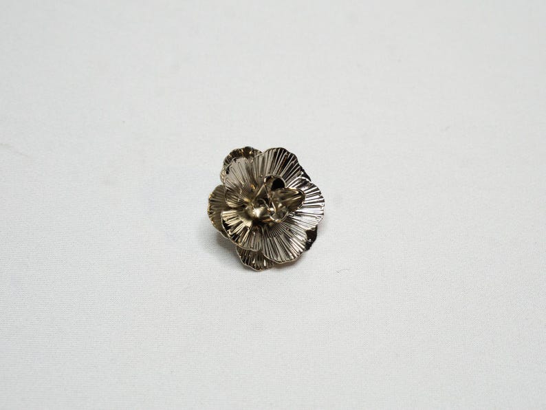 May include: A silver-coloured metal flower brooch. The brooch features multiple layers of petals with a textured, ridged design. The flower is centered on a white background, highlighting its intricate details.