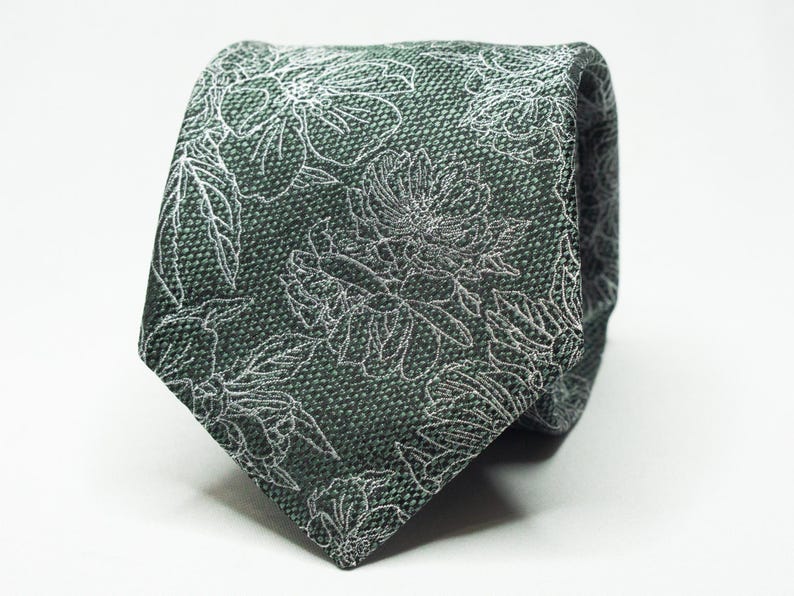 May include: A dark green tie with a floral pattern. The tie features a repeating design of white flowers and leaves. The fabric has a textured appearance, and the tie is knotted, showcasing its design.