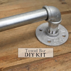 Puede incluir: Un kit de barra de toalla de acero galvanizado. La barra está hecha de tuberías y accesorios metálicos de estilo industrial. El kit incluye una base de brida redonda con agujeros pretaladrados para el montaje. El texto "Towel Bar DIY KIT" se muestra.
