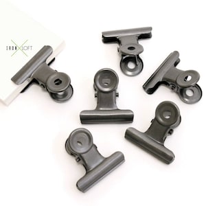 May include: Six black metal binder clips with a spring-loaded mechanism. The clips are designed to hold papers or other materials together.