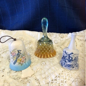May include: Three decorative bells, one is a blue and white ceramic bell with a windmill design, one is a clear glass bell with a yellow and orange iridescent finish, and one is a white ceramic bell with a bluebird and floral design.