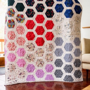 May include: A handmade quilt featuring a honeycomb pattern. The quilt showcases a variety of fabrics in shades of green, red, pink, purple, and blue. The quilt is displayed on a wooden floor.