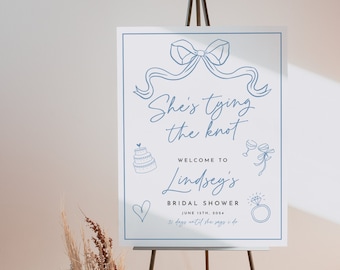 May include: A white sign with blue text and illustrations announcing a bridal shower for Lindsey. The sign reads "She's tying the knot" and "Welcome to Lindsey's Bridal Shower" with the date "June 15th, 2024" and the text "31 days until she says I do".