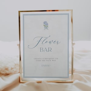 May include: A framed sign with the words "Flower Bar" in elegant script, with a small floral illustration above. The sign is in a gold frame and sits on a light-colored fabric. The text below reads "Create a bouquet to take on your way. Enjoy!"