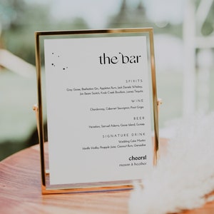 May include: A gold-framed sign with the text "the bar" in elegant script. The sign lists spirits, wine, beer, and signature drinks, including a Wedding Cake Martini. The sign is on a wooden tray with a blurred background.
