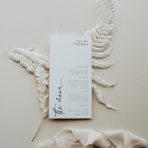 Peut inclure: Une carte de menu de mariage blanche avec du texte noir. Le menu comprend une bisque de crabe de Cornouailles, une salade de laitue Bibb locale, un filet mignon avec une sauce au vin rouge et un cheesecake aux baies fraîches. La carte est décorée de fougères séchées et d'un ruban blanc.