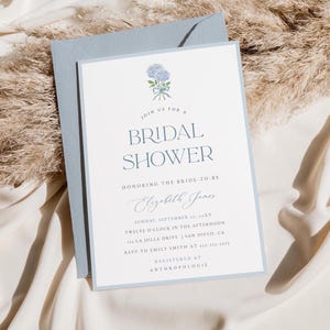 Puede incluir: Una invitación de color azul claro para una despedida de soltera con una ilustración de acuarela de un ramo de flores azules. La invitación dice "Join us for a Bridal Shower honoring the bride-to-be Elizabeth James. Sunday, September 1st, 10XX. Twelve o'clock in the afternoon. 133 La Jolla Drive | San Diego, CA. RSVP to Emily Smith at 135-555-5555. Registered at Anthropologie."